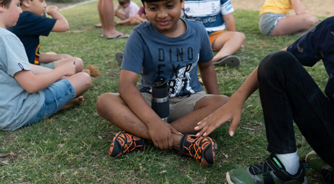 small indian boy sitting cross legged on the grass smiling at a fire he just learnt to make at edenbrook estate in parkhurst